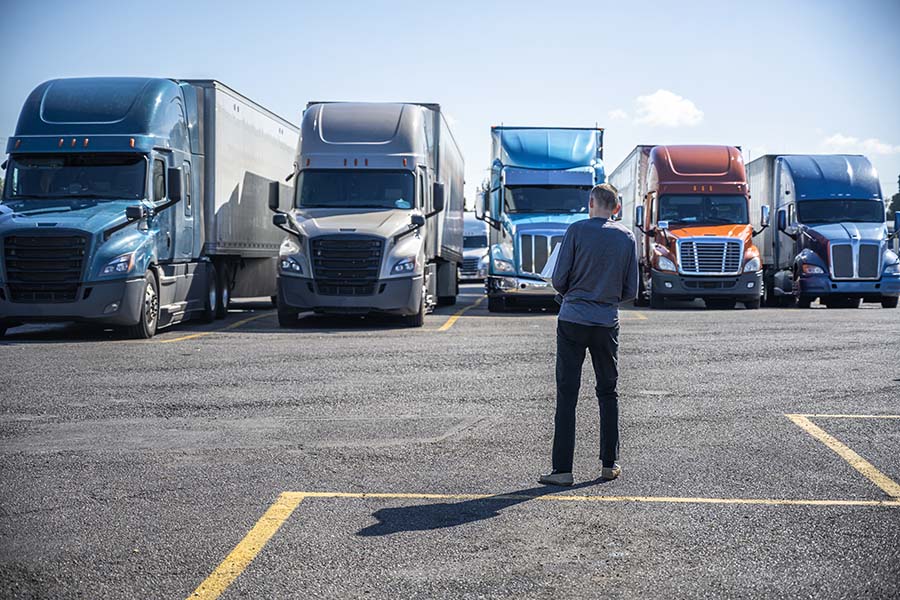Business Insurance - Rear View of a Man Standing in a Parking Lot Looking at a Row of Different Sized and Colored Tractor Trailer Trucks